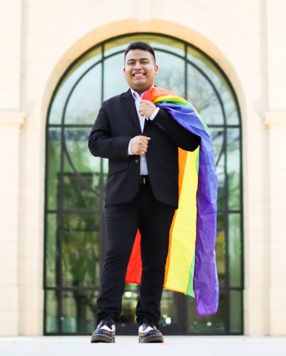 Photo of David Muñoz-Sarabia standing in front of a UNT building with a pride flag draped around their shoulders.
