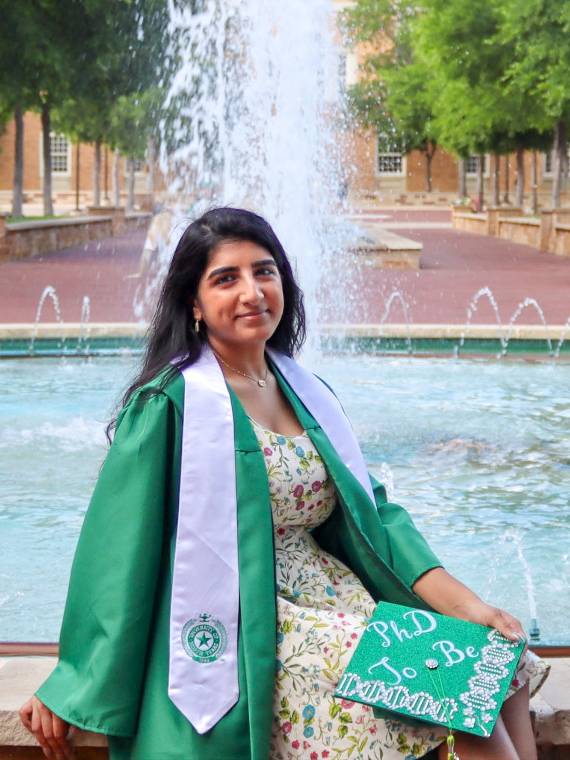 Kiran Daulla sits on the UNT fountain in commencement robes.