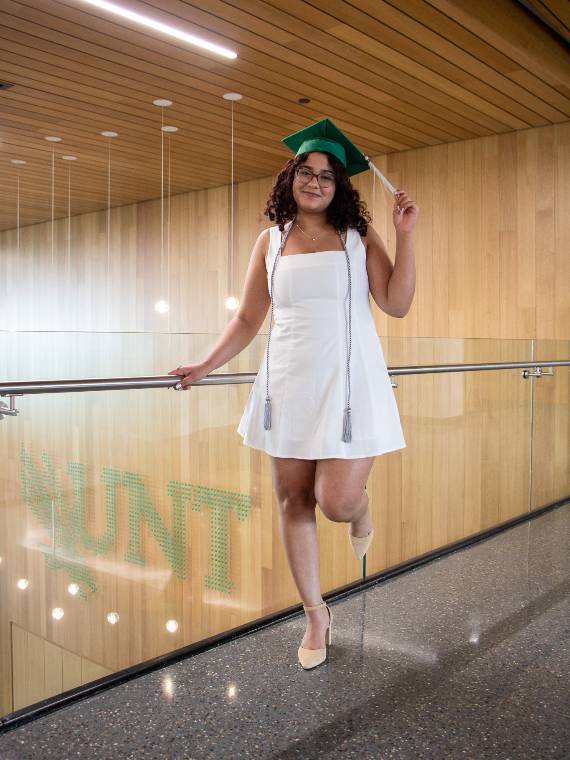 Nema Kolta poses on a balcony wearing her grad cap with a UNT installation in the background.