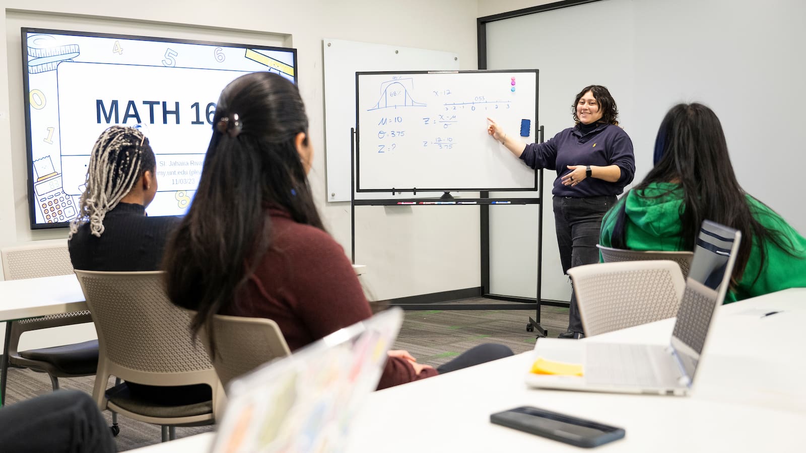 An instructor discusses math topics on a whiteboard with a classroom of young adult students