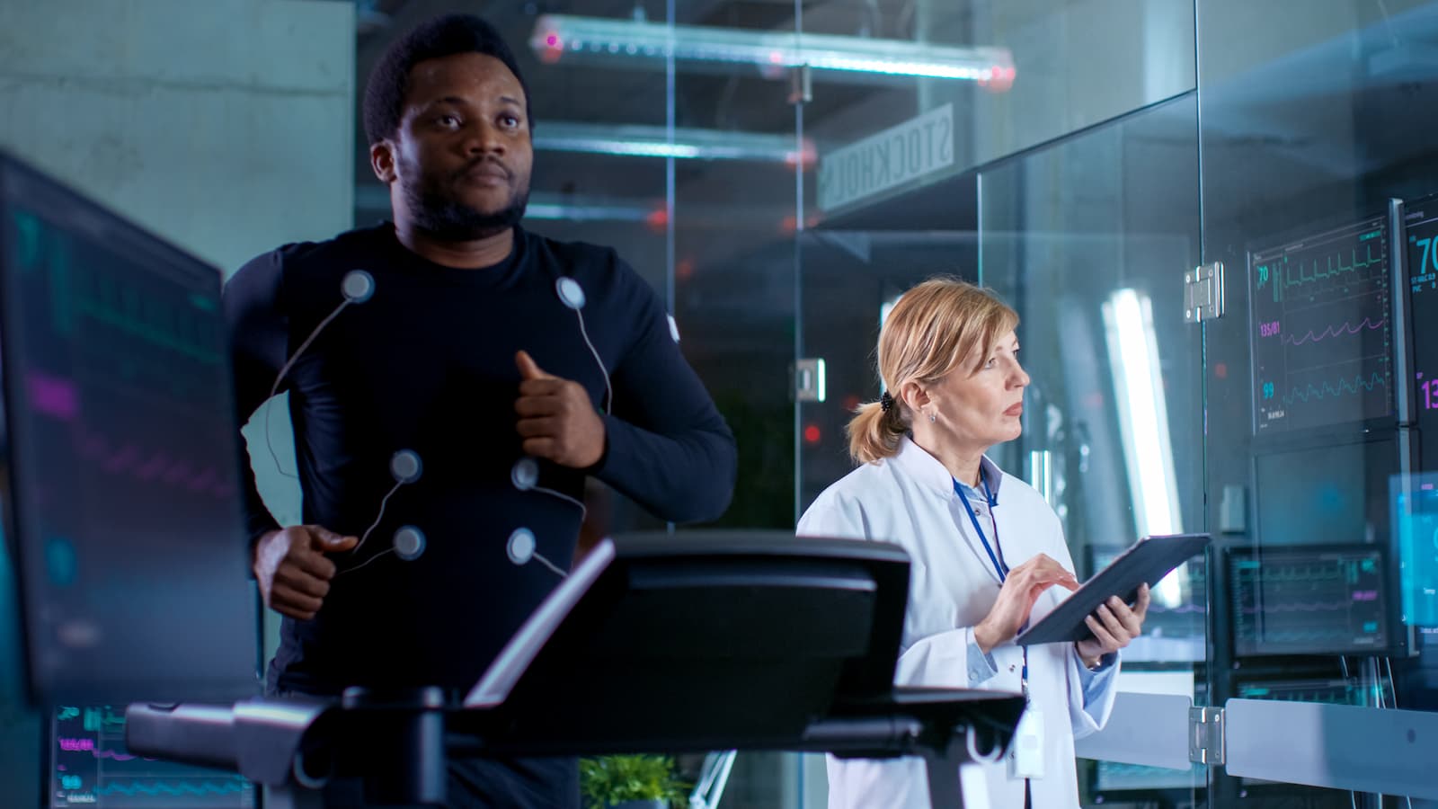 A man with sensors attached to his torso runs on a treadmill while a woman in a white lab coat looks at test results