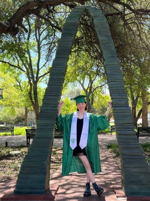 Alessandra Murrey poses in green commencement regalia under the UNT book arch sculpture.