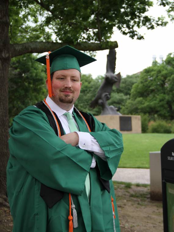 Jonathan Stewart posing in his commencement regalia with UNT's flying eagle statue in the background.