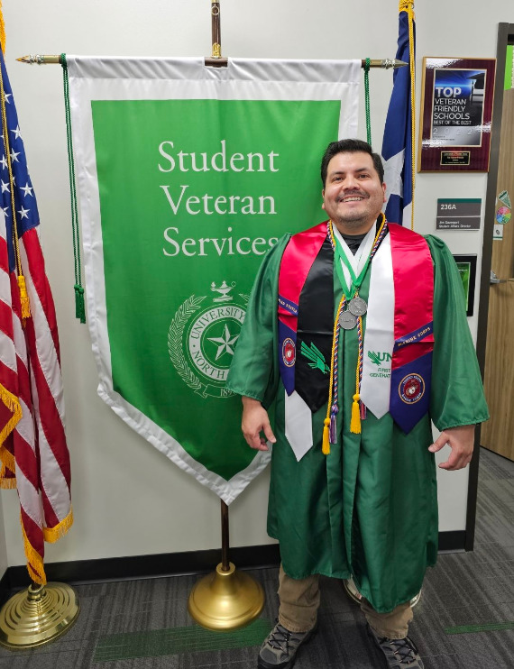 Maria Pena poses in front of a UNT Veterans flag wearing graduation regalia.