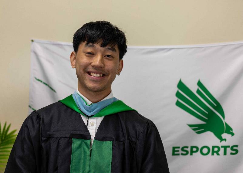 Vi Tran poses in commencement regalia in front of the UNT Esports flag