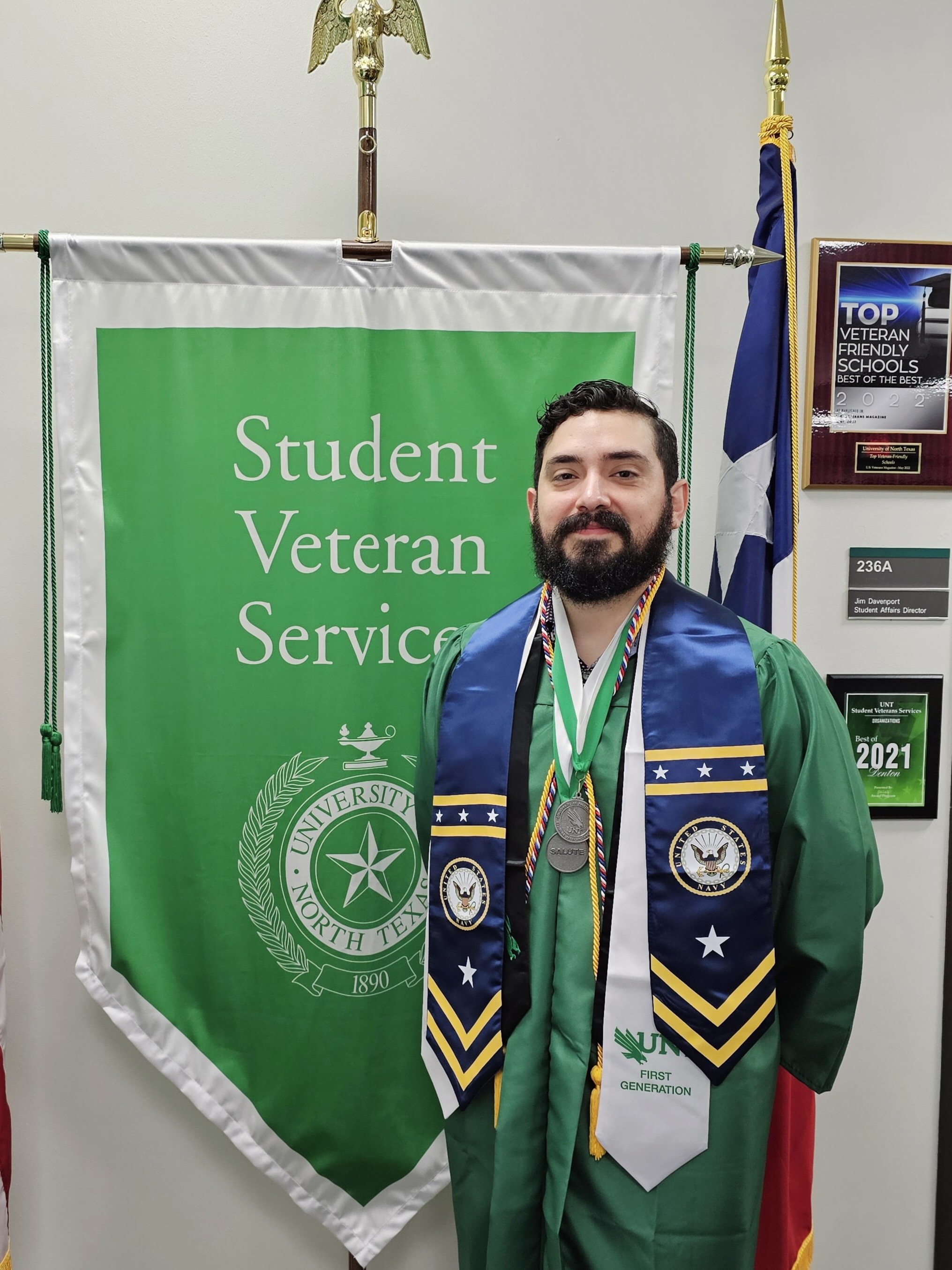 Walter Benitez wears his commencement regalia and stands in front of the UNT student veterans' flag