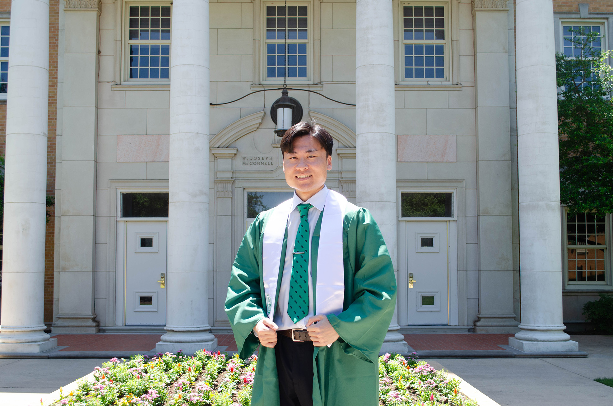 Yoshiki poses in commencement robe and green diving eagle tie in front of the UNT Administration Building