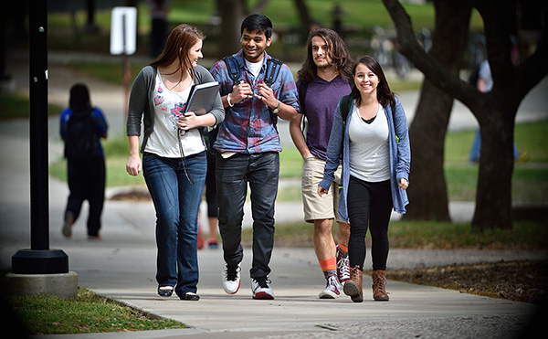 Four students holding books and wearing backpacks walking on a path.