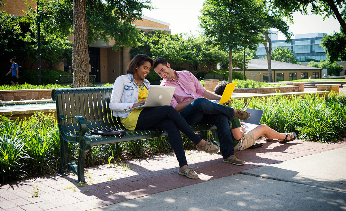 Two students looking at a laptop as they sit outside on a bench.
