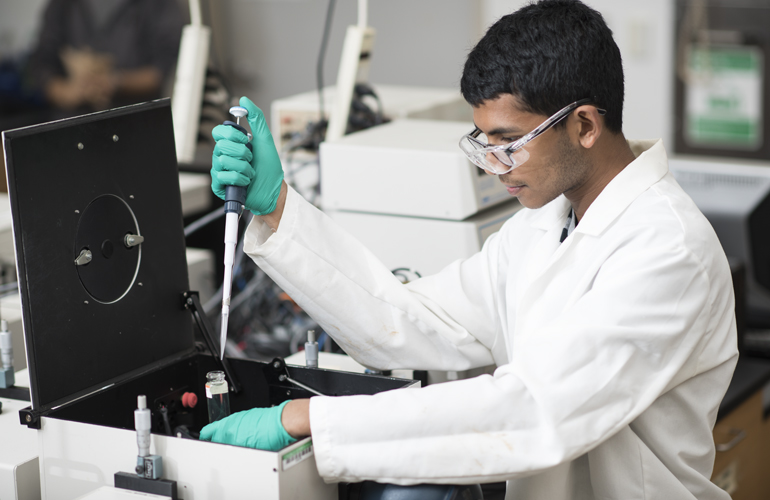 Student in a lab coat, with googles on, mixing chemicals.