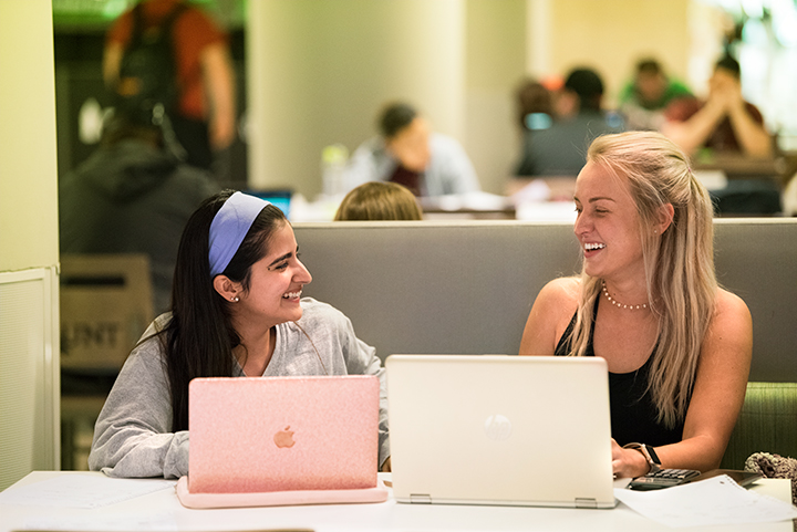 Two students talking to each other at the library with their laptops open. 